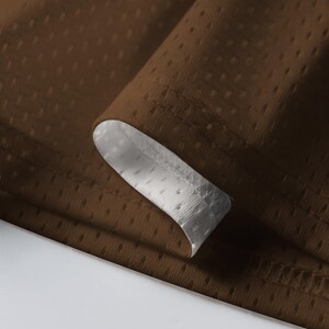 Puede incluir: Primer plano de tela marr&oacute;n con textura perforada. La tela est&aacute; doblada, revelando un forro blanco perforado. Los bordes est&aacute;n cuidadosamente cosidos, resaltando el contraste entre los materiales marr&oacute;n y blanco. La imagen muestra la textura y construcci&oacute;n del textil.