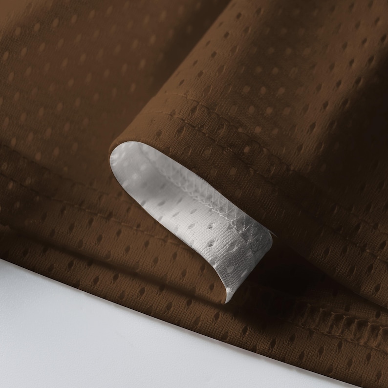 Puede incluir: Primer plano de tela marr&oacute;n con textura perforada. La tela est&aacute; doblada, revelando un forro blanco perforado. Los bordes est&aacute;n cuidadosamente cosidos, resaltando el contraste entre los materiales marr&oacute;n y blanco. La imagen muestra la textura y construcci&oacute;n del textil.