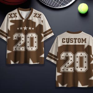 Puede incluir: Una camiseta deportiva marr&oacute;n y crema con el n&uacute;mero 20 y la palabra "CUSTOM" en la espalda. La camiseta tiene cuello en V y detalles de estrellas. La camiseta se muestra con una pelota de baloncesto, una raqueta de tenis y una pelota de tenis.