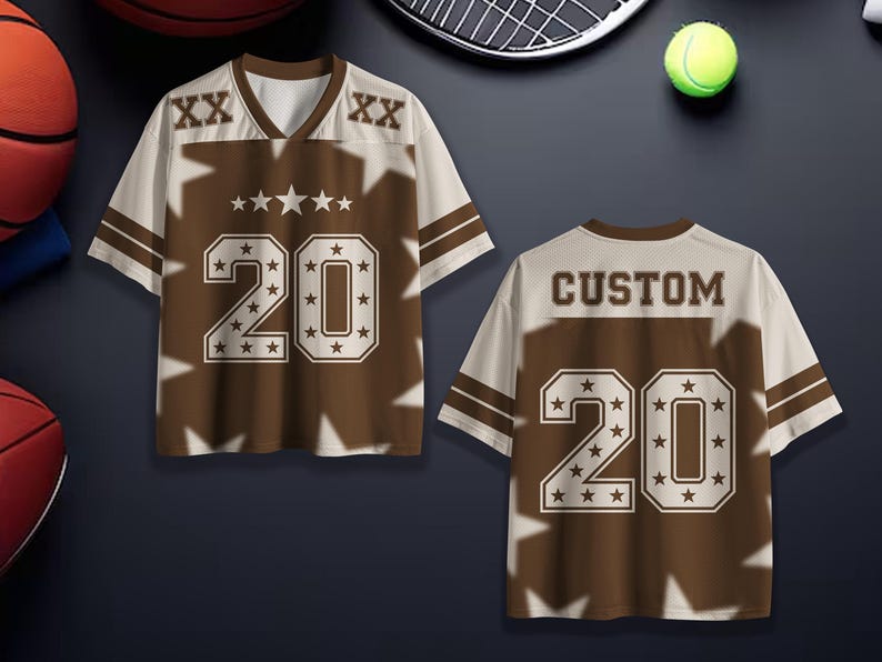 Puede incluir: Una camiseta deportiva marr&oacute;n y crema con el n&uacute;mero 20 y la palabra "CUSTOM" en la espalda. La camiseta tiene cuello en V y detalles de estrellas. La camiseta se muestra con una pelota de baloncesto, una raqueta de tenis y una pelota de tenis.
