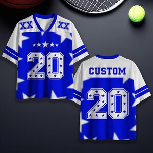 Puede incluir: Una camiseta deportiva azul y blanca con el n&uacute;mero 20 y detalles de estrellas. La camiseta tiene cuello en V y la palabra "CUSTOM" en la espalda. Se muestra con equipamiento deportivo de fondo.