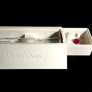 May include: A white box with the embossed text "Medha's Ants" contains a clear glass tube with a label and a small colony of ants. A smaller compartment holds two cylindrical objects, one red and one white.