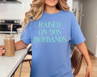 May include: A light blue t-shirt with the text "RAISED ON 90S BOYBANDS" in a light green color. The shirt is a short-sleeved, casual style.