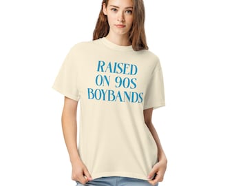 May include: Cream-colored t-shirt with the text "RAISED ON 90S BOYBANDS" in blue. The shirt has short sleeves and a relaxed fit. The model is wearing blue jeans.