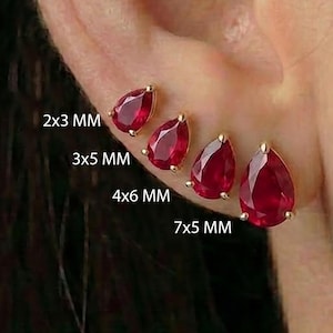 Puede incluir: Cuatro pendientes de botón con piedras preciosas rojas en forma de pera, dispuestos en una fila vertical en el lóbulo de la oreja. Los tamaños indicados son 2x3 mm, 3x5 mm, 4x6 mm y 7x5 mm. Cada piedra está engastada en metal dorado.
