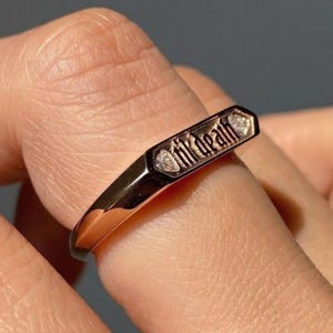 May include: A rose gold ring inscribed with "til death" in a gothic font. The ring features a rectangular top with two small diamond accents. The ring is on a finger.