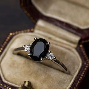 Op de afbeelding: Een zilveren ring met een grote ovale zwarte edelsteen, geflankeerd door twee kleinere witte edelstenen, gepresenteerd in een vintage bordeauxrode sieradendoos met een crèmekleurig interieur. De ring is het middelpunt.