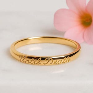 以下が含まれることがあります： 「Until Death」の文字が刻まれたゴールドカラーのリング。白い大理石の表面に置かれ、背景にはピンクの花があります。リングはシンプルなバンドデザインです。