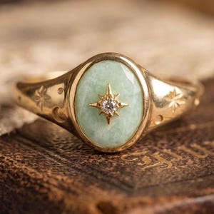 May include: Gold ring featuring an oval, pale green stone and a starburst design with a small diamond. The ring has a detailed band with star and crescent moon engravings.