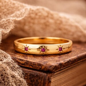 Op de afbeelding: Een gouden ring met drie stervormige ontwerpen, elk met een kleine, ronde, rode edelsteen. De ring ligt op een vintage boek en gedrapeerde stof, wat een warme, antieke esthetiek creëert.