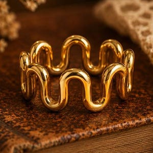 Puede incluir: Un anillo de tono dorado que presenta un diseño ondulado o serpentino continuo y repetitivo. La banda está construida con un alambre de metal liso y pulido que se curva hacia arriba y hacia abajo en un patrón constante.