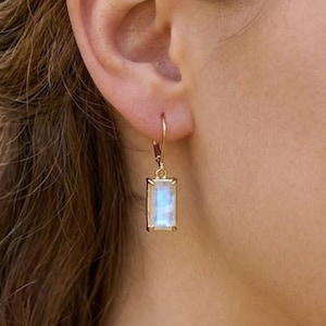May include: A gold drop earring featuring a rectangular, iridescent moonstone set in a simple metal frame. The earring hangs from a thin gold hook, showcasing a smooth, polished finish and a soft, glowing light blue sheen on the stone surface.