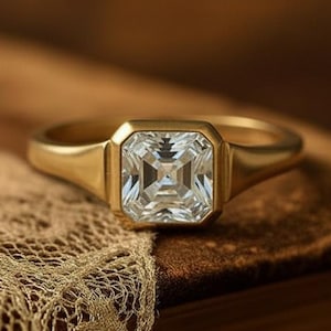 May include: A gold ring featuring a square-cut, clear gemstone set in a bezel setting. The band is smooth and polished, showcasing a clean, metallic finish that contrasts with the faceted, reflective surface of the central stone.