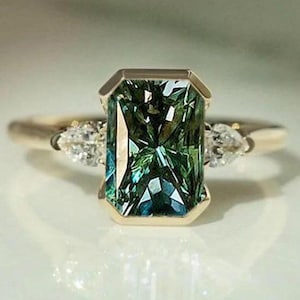 Puede incluir: Un anillo de oro con una gran piedra preciosa verde rectangular de corte esmeralda en un engaste de bisel. La piedra central está flanqueada por dos piedras claras más pequeñas en forma de pera, sobre una banda de oro pulido.