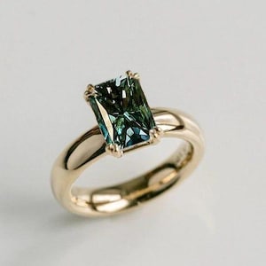 May include: A gold ring featuring a rectangular emerald-cut teal green gemstone set in a four-prong setting. The band is smooth, polished, and rounded, showcasing a high-shine metallic finish that complements the deep, multi-faceted green stone.