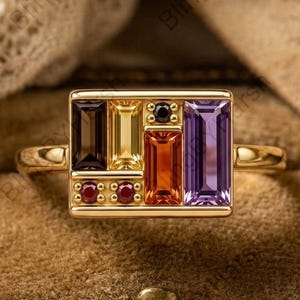 May include: A gold ring featuring a rectangular face set with a vertical arrangement of multi-colored gemstones, including a dark brown, yellow, orange, and purple baguette-cut stone, alongside two small round red stones and one black stone.