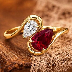 Könnte beinhalten: Ein Bypass-Ring aus poliertem Gelbgold mit zwei birnenförmigen Edelsteinen. Ein Stein ist ein klarer, facettierter Diamant, der andere ein tiefroter, facettierter Rubin. Die Ringschiene schwingt sich in einem fließenden Design um die Steine.