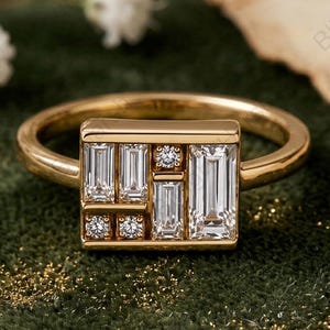 Pode incluir: Um anel de ouro com um engaste retangular apresentando uma disposição geométrica de várias pedras claras de corte baguette e redondo. O aro é liso e polido, exibindo um design limpo e estruturado com um acabamento metálico brilhante.