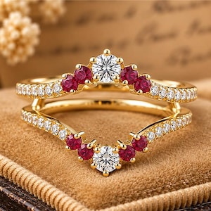 May include: A gold ring set featuring two chevron-shaped bands. The top band has a central clear round stone flanked by four red round stones and pavé-set clear stones. The bottom band mirrors this design with red and clear stones.