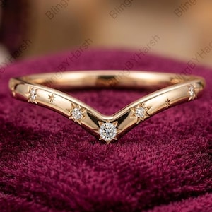 May include: A V-shaped ring crafted from polished rose gold, featuring a chevron silhouette. The band is adorned with five small, clear, round-cut stones, each set within a star-shaped engraving, creating a celestial pattern across the front.
