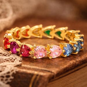 May include: A gold band ring featuring a continuous row of heart-shaped gemstones in various colors, including red, pink, blue, and green. Each stone is held in a prong setting, creating a vibrant, multi-colored circular design.