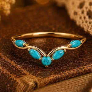 May include: A gold-colored ring with a V-shaped design, adorned with five oval-shaped turquoise stones. The ring is set on a brown leather-bound book, with a blurred background and a decorative lace cloth.