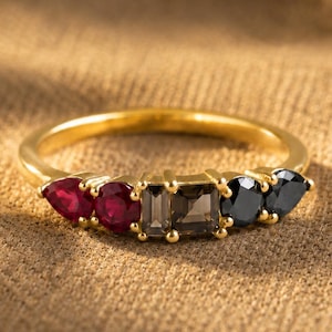 May include: A gold ring featuring a row of five multi-shaped gemstones. The arrangement includes two pear-cut red stones, two rectangular-cut smoky brown stones, and two round-cut black stones set in a polished gold band with prong settings.
