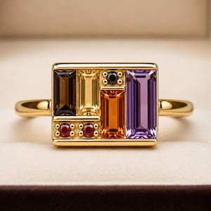 May include: A gold ring with a rectangular face, set with a variety of coloured gemstones. The gemstones are brown, yellow, orange, purple, and red. The ring is in a gold band and is presented in a brown jewellery box.