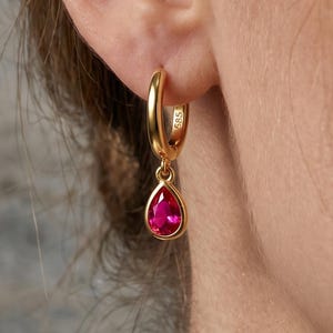 May include: A gold hoop earring featuring a teardrop-shaped fuchsia gemstone pendant. The hoop is stamped with the number 585, indicating its gold content. The earring is suspended from an earlobe, showcasing the polished metal and vibrant stone.