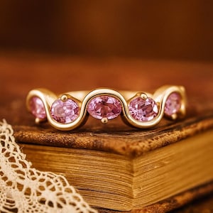 Peut inclure: Une bague en or ornée de cinq pierres roses ovales. Les pierres sont serties dans un motif ondulé. La bague est présentée sur un livre ancien, avec un détail de dentelle au premier plan. C'est un bijou.