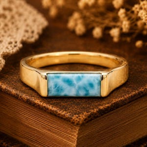 May include: A gold ring featuring a rectangular, horizontally set, light blue Larimar gemstone with a marbled pattern. The band is smooth and polished, showcasing a clean, metallic finish that contrasts with the textured, sky-blue stone inlay.
