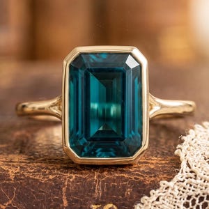Pode incluir: Um anel de ouro com uma grande pedra preciosa retangular de cor verde-azulado. O anel está sobre um livro antigo com letras douradas e uma delicada toalha de renda. A pedra preciosa é o ponto focal, mostrando sua cor vibrante e corte.