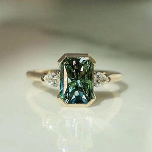 Puede incluir: Anillo de oro con una piedra verde rectangular con talla esmeralda y matices azules. Dos piedras claras más pequeñas en forma de pera flanquean la piedra central. El anillo tiene una banda pulida.