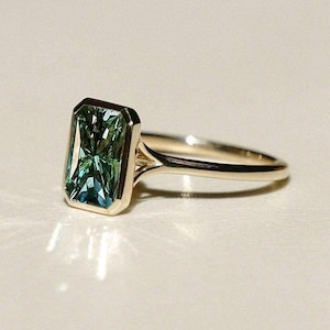 May include: A gold ring with a rectangular, emerald-cut gemstone displaying green and blue tones. The gemstone is set in a bezel setting, and the ring has a slender band.