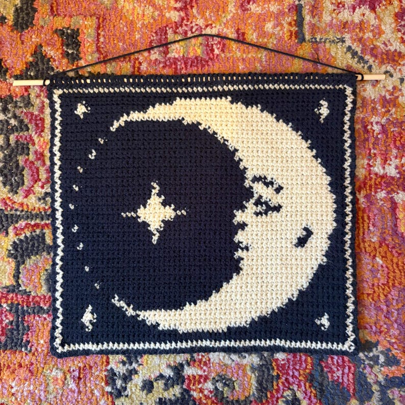 May include: A navy blue, square crochet wall hanging with a cream-coloured crescent moon and stars design. It has a white border and is suspended from a wooden dowel, perfect for a bedroom or living room.