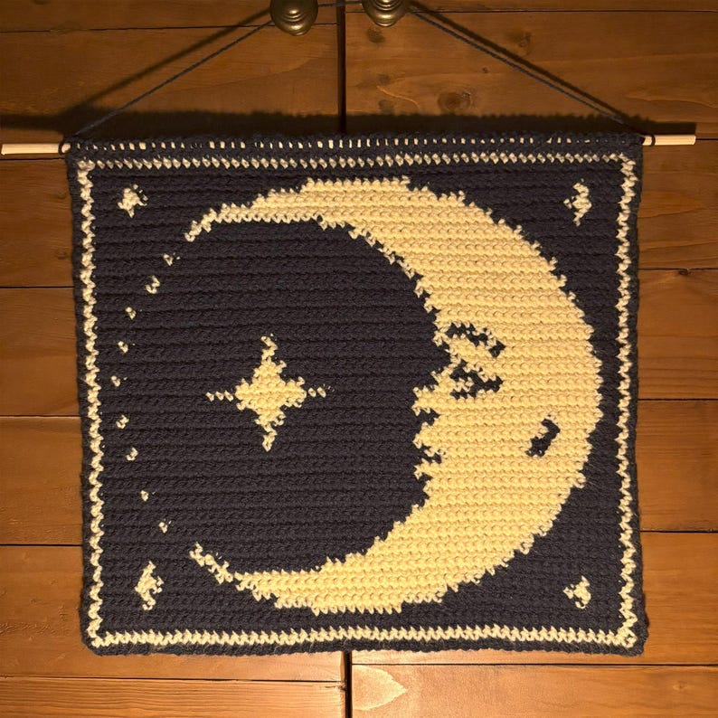 May include: A navy blue crocheted wall hanging with a cream-coloured crescent moon and star design. The rectangular piece is finished with a cream border and has a wooden dowel at the top for hanging.
