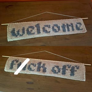 Op de afbeelding: Een gehaakte banner in beige met het woord "welcome" in donkerblauwe letters. De banner hangt aan een houten deuvel met een touwtje. De tweede banner zegt "fuck off".
