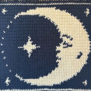 May include: A crocheted item with a dark blue background, displaying a cream-colored crescent moon and stars. The piece has a textured feel, with the moon and stars standing out against the dark blue. The design evokes a night sky, with detailed crochet work.