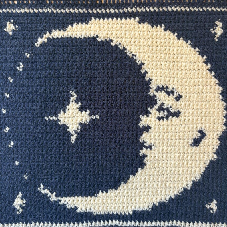 May include: A crocheted item with a dark blue background, displaying a cream-colored crescent moon and stars. The piece has a textured feel, with the moon and stars standing out against the dark blue. The design evokes a night sky, with detailed crochet work.