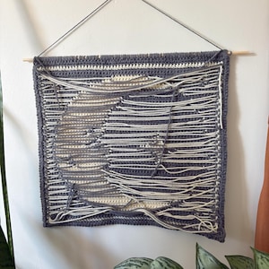 May include: A handmade, square wall hanging with a unique design. The piece features a dark grey crocheted border and a central design in cream and grey, with horizontal white yarn strands. It is suspended from a wooden dowel with a dark grey string.