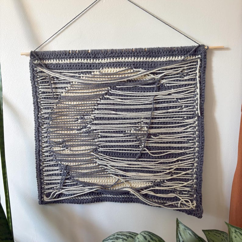 May include: A handmade, square wall hanging with a unique design. The piece features a dark grey crocheted border and a central design in cream and grey, with horizontal white yarn strands. It is suspended from a wooden dowel with a dark grey string.