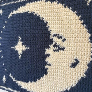 May include: Close-up of a crocheted blanket with a cream-coloured crescent moon and stars against a navy blue backdrop. The blanket is made with a textured stitch, showcasing the detailed craftsmanship.