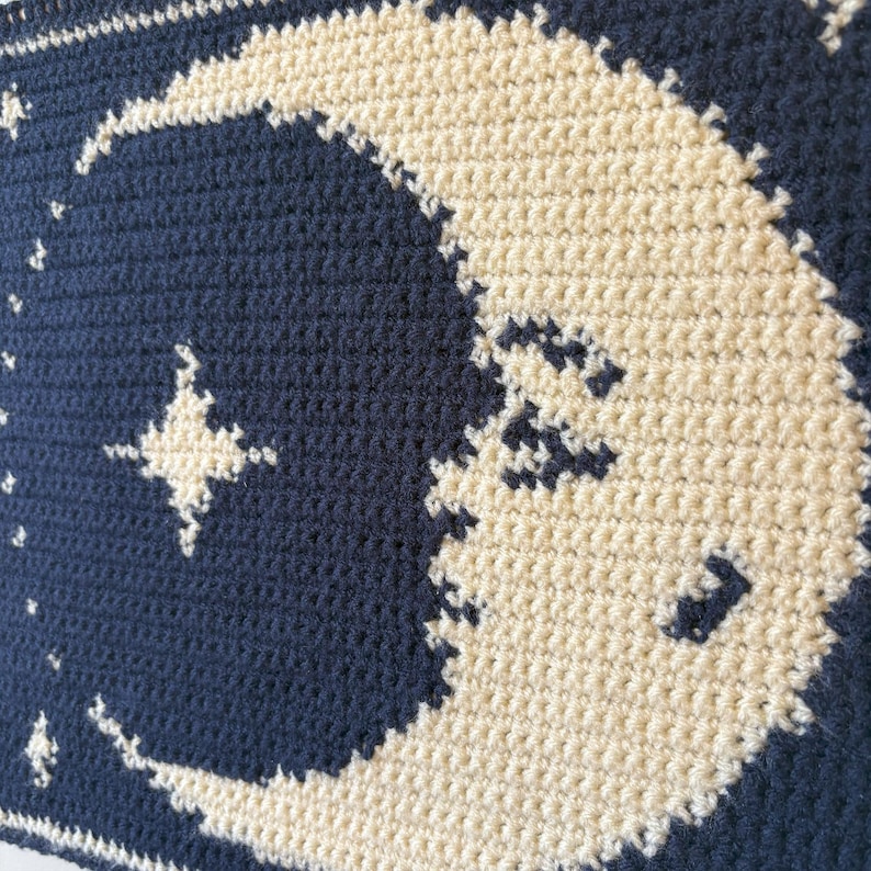 May include: Close-up of a crocheted blanket with a cream-coloured crescent moon and stars against a navy blue backdrop. The blanket is made with a textured stitch, showcasing the detailed craftsmanship.