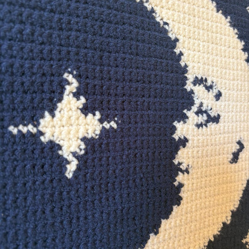 May include: Close-up of a navy blue and cream-coloured crocheted cushion. The cushion features a repeating pattern of a star and a crescent moon. The star and moon are cream-coloured and the background is navy blue.