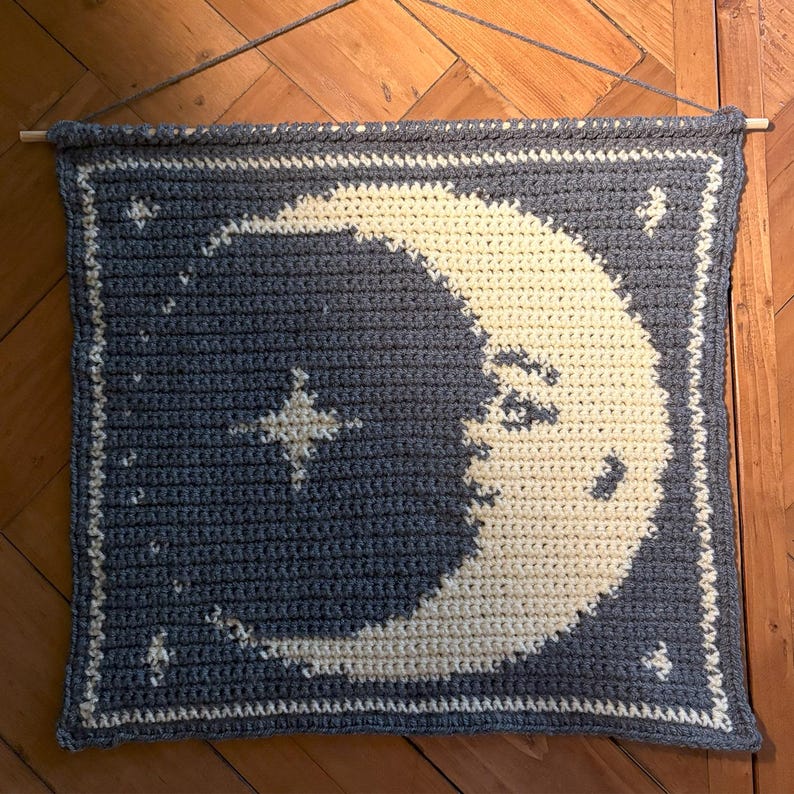 May include: A crocheted wall hanging with a crescent moon and stars design. The moon and stars are cream-coloured against a dark blue backdrop. The wall hanging is rectangular and suspended from a wooden dowel.