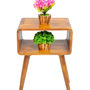 May include: A wooden side table with a two-tiered design. The table features a warm, natural wood finish and has a curved, open shelf. Two small potted plants, one with pink flowers and the other with green foliage, sit on the table.