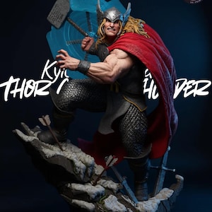 May include: A detailed statue of Thor, the Marvel Comics superhero, wielding his hammer and a blue energy shield. He wears a silver helmet, chainmail, and a red cape. The statue is on a rocky base with arrows and swords.