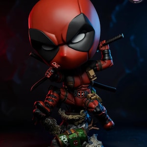 Puede incluir: Figura de Deadpool rojo y negro en una pose dinámica, sosteniendo armas. La figura está sobre una base rocosa con un contenedor verde y marrón. El texto "KyleVerseStudio" está en la parte inferior.