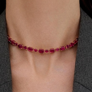 Puede incluir: Un collar de oro con una serie de piedras preciosas rectangulares de color rojo intenso. El collar se lleva alrededor del cuello, con las piedras preciosas dispuestas en una línea continua. El fondo es una blazer gris.