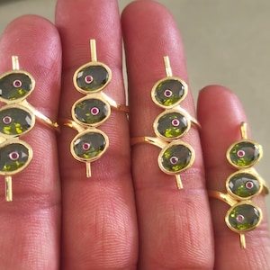Puede incluir: Cuatro anillos de oro, cada uno con tres piedras ovaladas de color verde oliva alineadas verticalmente. Cada piedra está acentuada con una pequeña piedra preciosa circular de color rosa intenso en el centro. Tienen una banda de oro pulido y una barra.